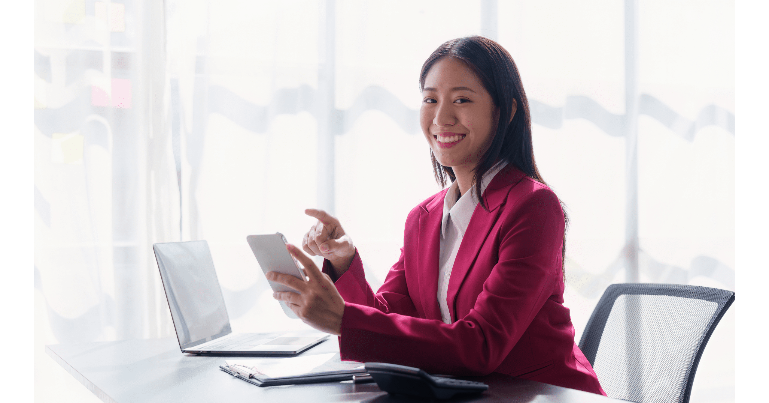 Smiling businesswoman in a red blazer using a smartphone at her desk with a laptop and documents.