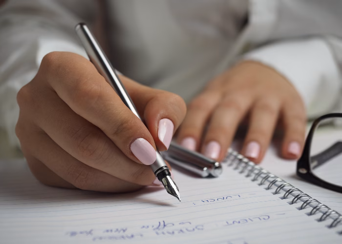 Close-up of a woman writing notes in a spiral notebook with a fountain pen, next to eyeglasses.