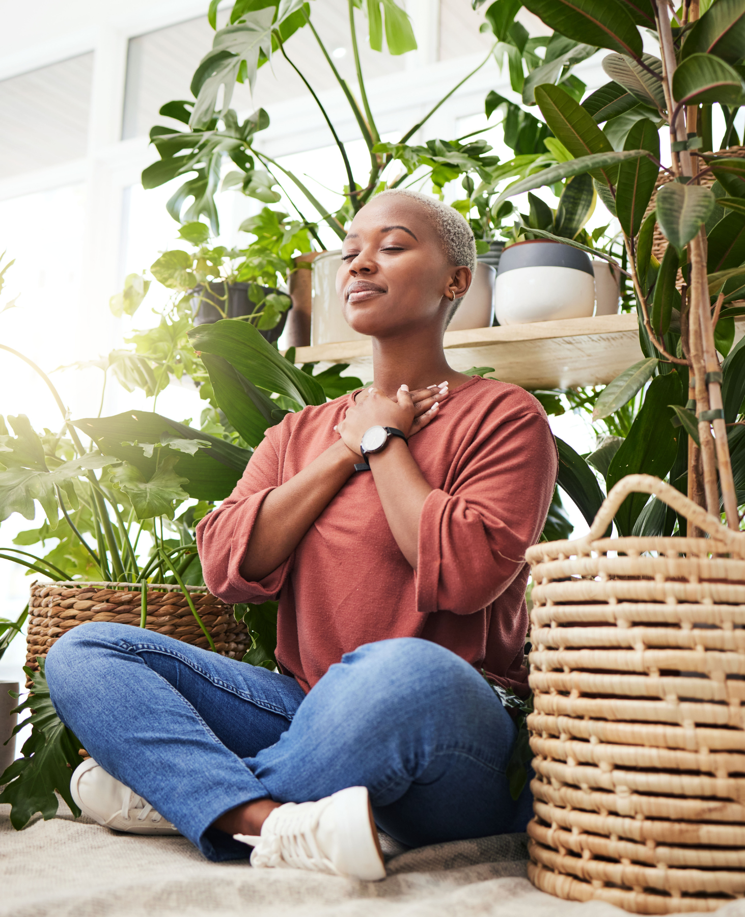Peaceful woman sitting cross-legged among indoor plants with eyes closed and hands on heart, embracing mindfulness and gratitude.